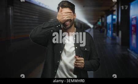Hispanic man holding beer bottle covers eyes with hand on train platform inside building; embarrassment late commute. Stock Photo