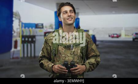 Soldier holding binoculars and dog tags, smiling and looking upward at pumps on a street; duty vigilance. Stock Photo