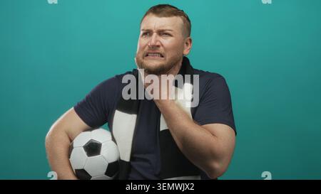 Man holding soccer ball while wearing black and white fan scarf, clutching throat with pained expression in teal studio backdrop; pain. Stock Photo