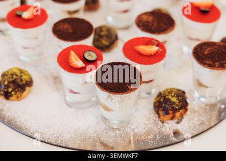 Tartlet desserts in clear glasses with red fruit topping. Cocoa powder dusted over creamy base in each glass. Sweet treats arranged on reflective silv Stock Photo