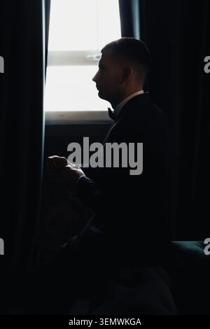 Silhouetted man sits by window, gazing outward. Hands folded, posture still, quiet contemplation. Dark curtains frame bright window light behind. Mood Stock Photo