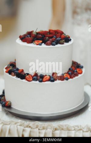 White tiered cake adorned with fresh berries. Strawberries, blueberries, and raspberries decorate the cake. Soft lighting enhances the cake's delicate Stock Photo