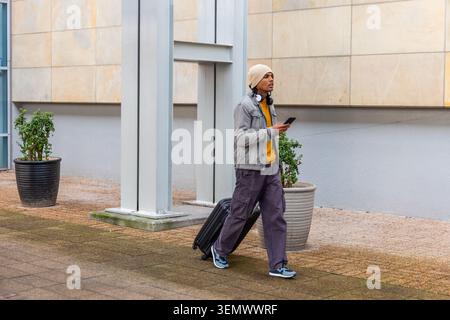 Asian man walking plaza by tiled wall pulling suitcase holding phone wearing headset yellow hoodie Stock Photo