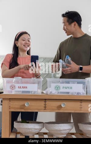 Asian couple sorting waste at kitchen island tapping phone and holding bottles by GLASS or PAPER bin Stock Photo
