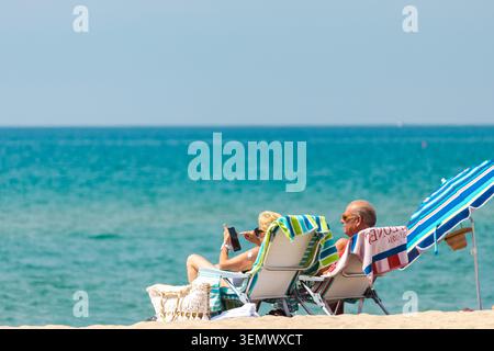 Sant Salvador, Spain, 7 June 2026: Senior relaxing in beach chairs on Mediterranean shoreline and using smartphone Stock Photo