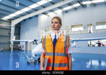 Woman wearing a safety vest and carrying a laptop, standing confidently in an aircraft hangar with light airplanes, symbolizing career, aerospace, and Stock Photo