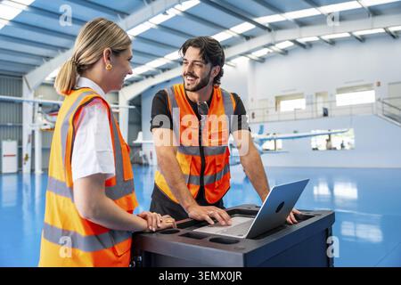 Two aviation engineers wearing high visibility vests discussing aircraft maintenance, working on a laptop at a portable workstation in an airplane han Stock Photo