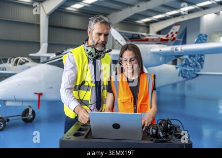 Two aviation engineers in high visibility vests and safety glasses review laptop data and discuss maintenance and inspection plans inside a hangar wit Stock Photo