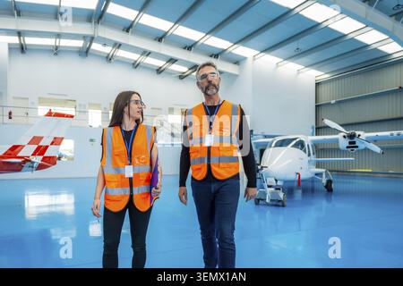 Aviation engineers wearing safety vests and glasses. Walking and discussing in an airplane hangar with light aircraft undergoing maintenance. Represen Stock Photo