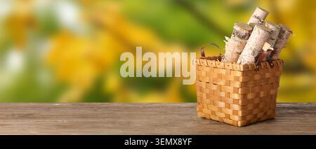 Birch firewood logs in woven basket on rustic wooden table with blurred green yellow background Stock Photo