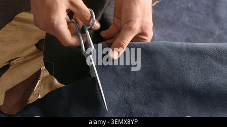 Craftsman cutting dark blue leather with scissors for handmade goods and upholstery projects, this process involves careful handling of materials for Stock Photo