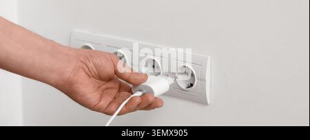 Hand plugging white charger into electrical outlet on white wall, power supply and device charging Stock Photo