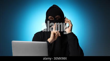 Cybercrime and data security: hacker in balaclava mask with laptop and smartphone, shushing gesture for secrecy, cybercrime and data security are crit Stock Photo