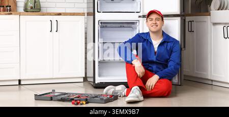 Smiling male appliance repair technician sitting on kitchen floor next to open refrigerator with tool kit, a tool kit is placed on the floor in front Stock Photo