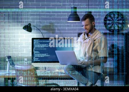 Smiling male programmer working on laptop and desktop computer with data overlay, software development and cybersecurity Stock Photo