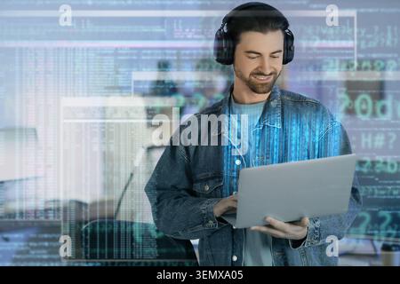Smiling male programmer in headphones using laptop with digital code overlay software development and cybersecurity, the man is focused on his work em Stock Photo