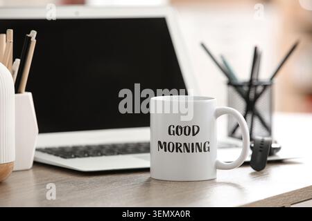Good morning coffee mug on wooden desk with laptop and office supplies, remote work and home office setup, office supplies including pens in a holder Stock Photo