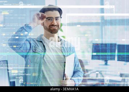 Smiling male software developer with glasses and laptop, programming code overlay, digital technology and innovation Stock Photo