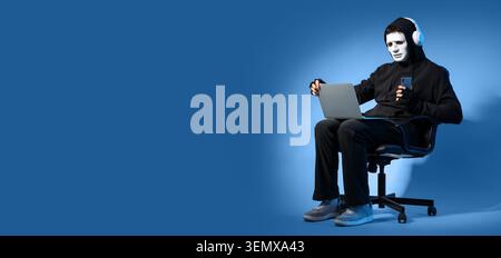 Cybersecurity threat: anonymous hacker in mask and hoodie using laptop and smartphone for data breach and online fraud, the individual engages in data Stock Photo