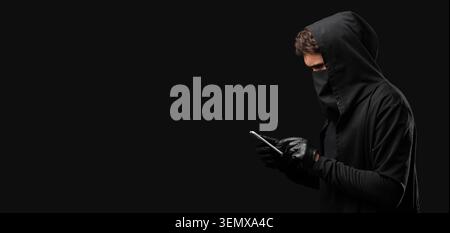 Cybercrime and data security with hooded hacker using smartphone on dark background, the individual wears gloves while interacting with the mobile dev Stock Photo