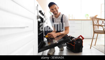 Professional appliance repairman fixing oven in modern kitchen, home maintenance and service concept, the skilled worker in uniform uses a screwdriver Stock Photo