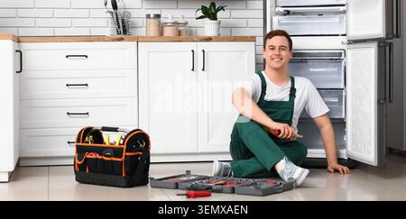 Smiling young male appliance repair technician in overalls sitting on kitchen floor with open refrigerator and tool kitchen Stock Photo