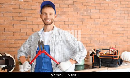 Smiling young repairman in blue overalls and cap holding bolt cutters with toolbox in kitchen, a toolbox sits on the kitchen counter in the background Stock Photo