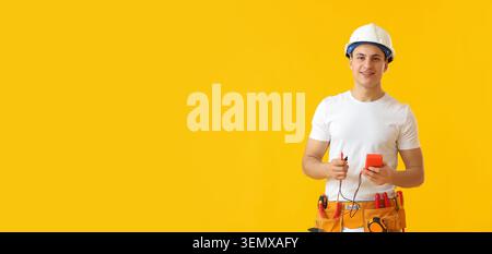 Smiling young electrician in hard hat and tool belt holding multimeter on yellow background with copy space Stock Photo
