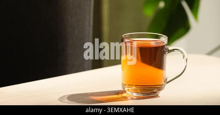 Clear glass mug with amber tea on wooden table, warm beverage for relaxation and comfort, natural light Stock Photo