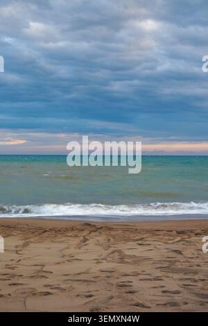 Waves crash on sandy beach under cloudy sky at sunset Stock Photo