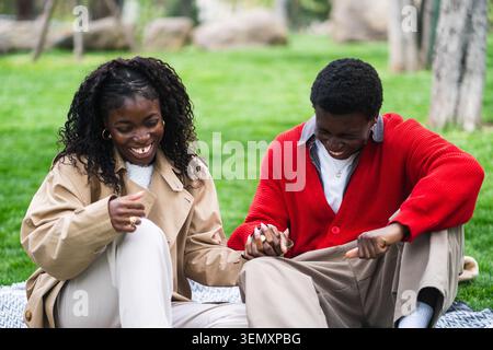Cheerful positive african american man and woman sitting on plaid in park laughing and joking Stock Photo