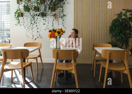 A woman is seated at a table in a bright cafe. She is holding a coffee and looking thoughtful. Sunflowers and other flowers are on the table. Empty ch Stock Photo