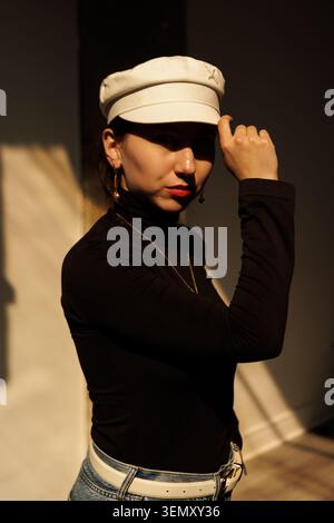 A woman stands indoors with a white hat and a black shirt. She touches her hat while casting a shadow. The light creates a warm atmosphere in the back Stock Photo