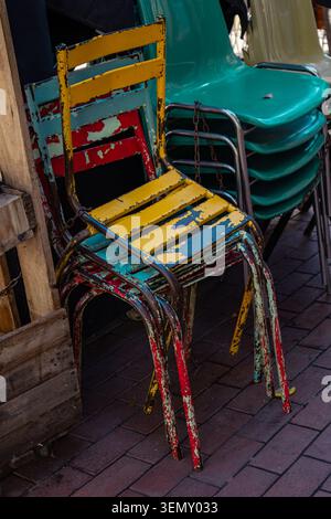 A collection of colorful weathered metal chairs and stacked plastic seats arranged on an outdoor café terrace. The worn paint, vibrant colors, and lay Stock Photo