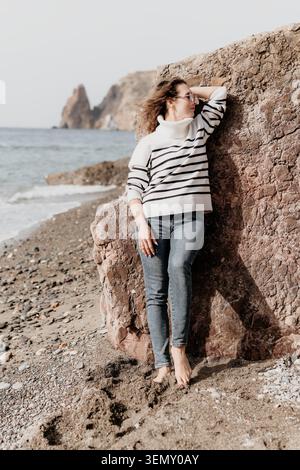 Woman Coast Relaxation Leaning against a large rock on a pebble beach with ocean views during a serene vacation Stock Photo