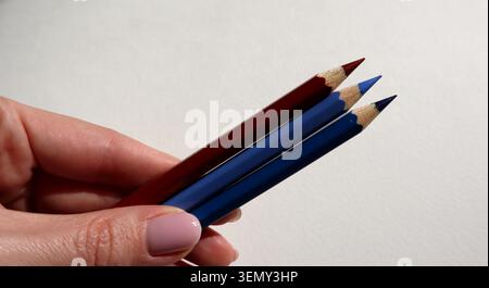 A hand holds two colored pencils, one red and one blue. The pencils have sharp tips and are placed on a plain background. This scene happens around midday with natural lighting. Stock Photo