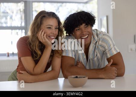 Couple leaning on white kitchen counter, smiling and wearing casual tops beside beige ceramic bowl Stock Photo