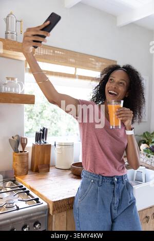 Smartphone resting next to glass of orange juice on wooden countertop in home kitchen with utensils Stock Photo