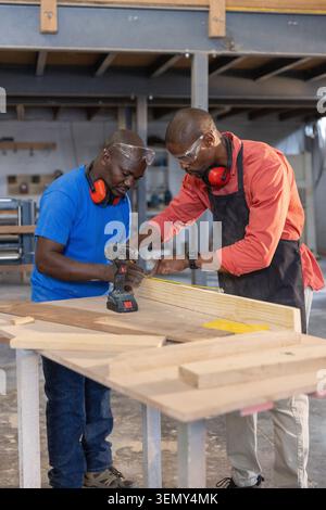 African American coworkers using tape measure and placing cordless drill on workbench at workshop Stock Photo