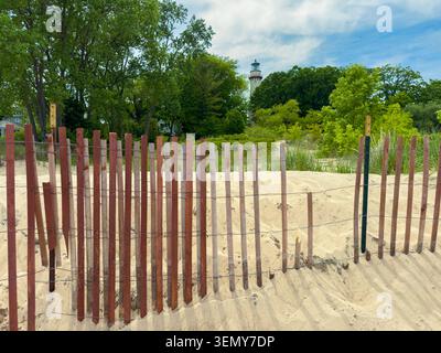 Fencing made of wood marks the edge of sandy dunes. A lighthouse is visible in the distance with green trees nearby under a blue sky with clouds. Stock Photo