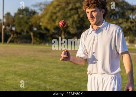 Man bowling on field wearing white cricket uniform, red leather cricket ball in air, copy space Stock Photo