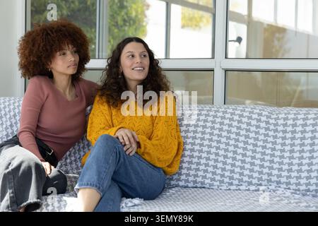 Diverse female friends sitting on patterned couch in living room with houndstooth throw, smiling Stock Photo