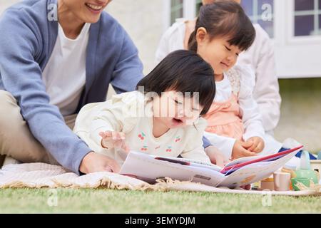 Japanese family parents and two daughters an infant and a preschooler reading a picture book on a blanket outdoors Stock Photo