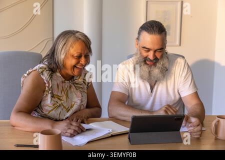 Senior couple laughing, reviewing notes at home table, in floral blouse and polo, using tablet Stock Photo