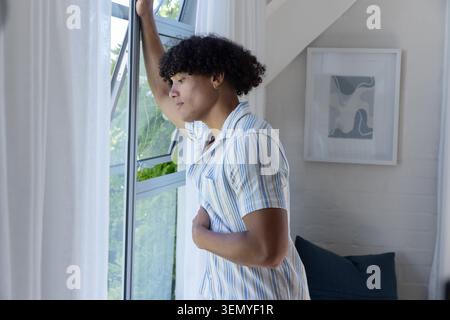 African American man in his 20s leaning by multi-pane window wearing striped shirt, gazing outside Stock Photo