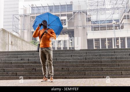 African American man standing on office steps in orange top holding blue umbrella using smartphone Stock Photo