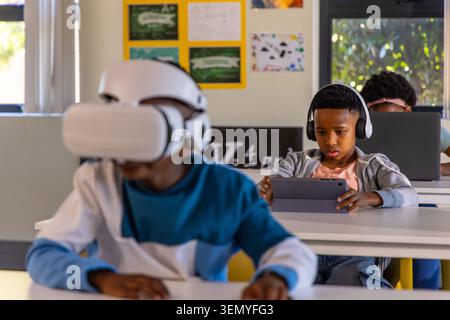 African American schoolchildren sitting at desks holding tablet, wearing headphones, VR headset Stock Photo