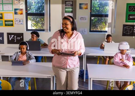 African American teacher with students holding tablet at front of classroom while using devices Stock Photo