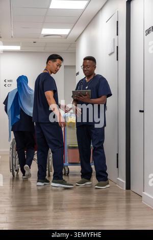 Diverse medical staff in navy scrubs consulting tablet and pushing wheelchair in hospital corridor Stock Photo