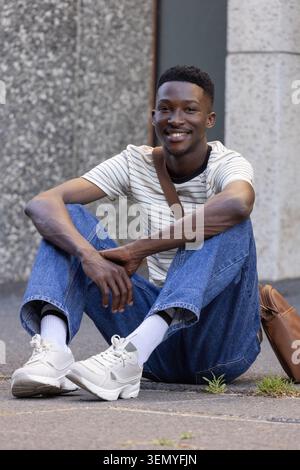 Adult African man sitting on pavement by concrete facade smiling, wearing striped T-shirt, backpack Stock Photo